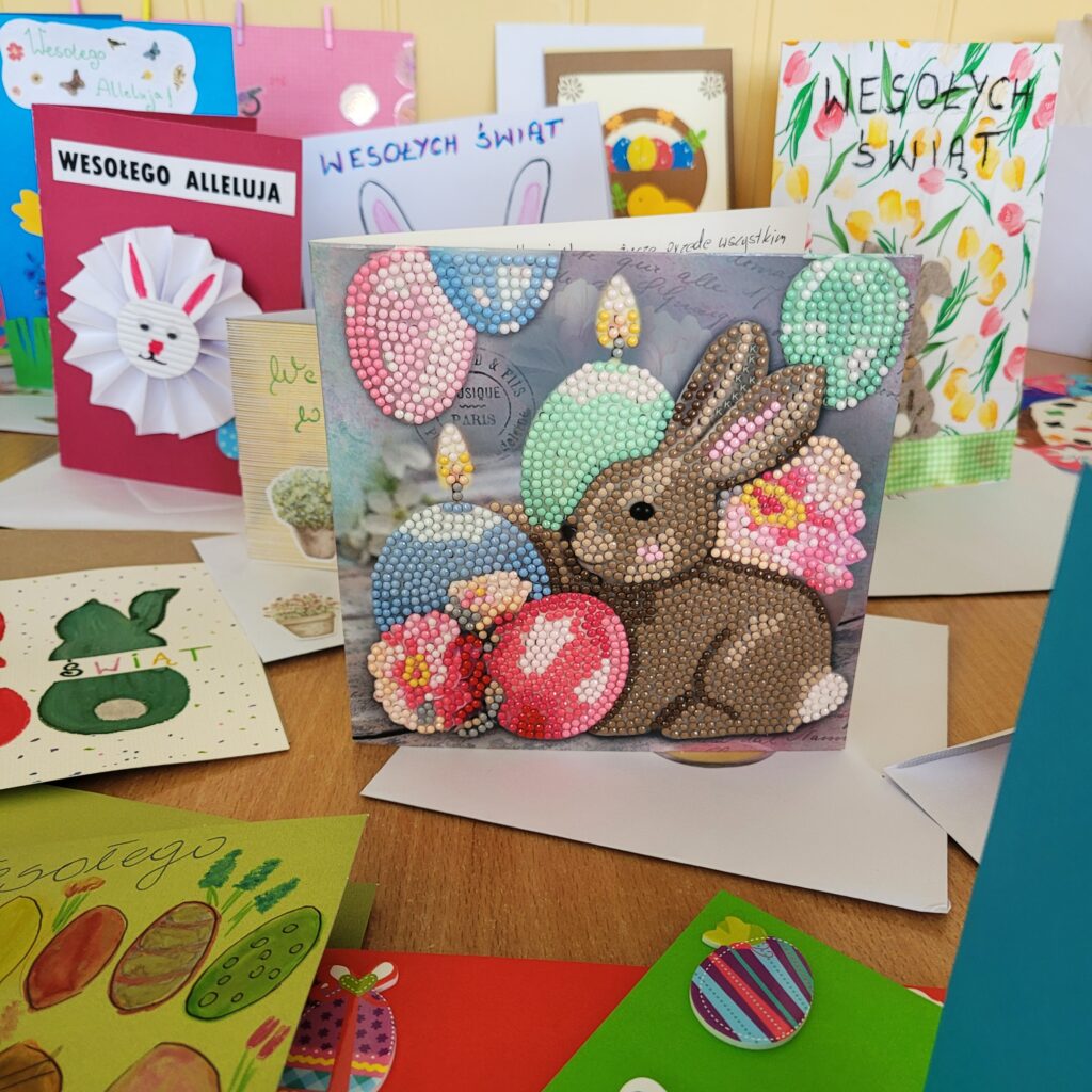 Wyeksponowana na tle innych kartek, kartka z zajączkiem i świeczkami w kształcie jajeczek, wykonana ciekawą techniką zwaną mozaiką diamentową.