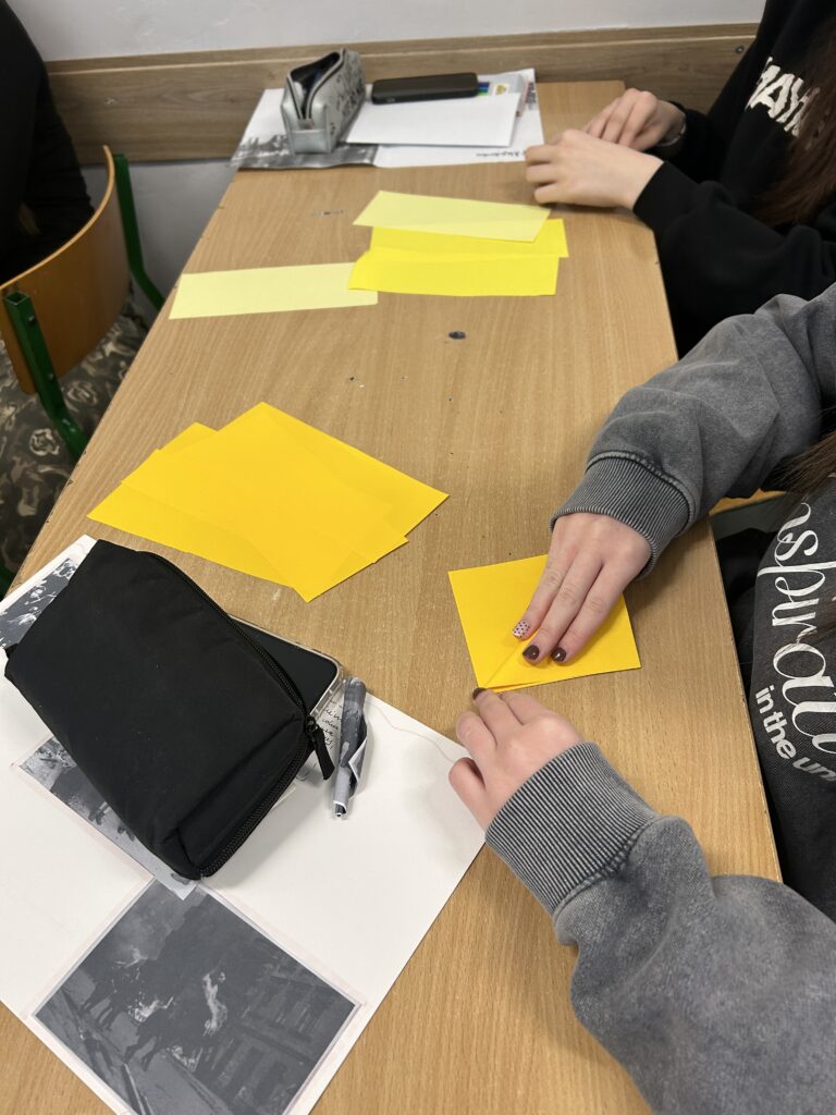 na ławce leżą różne kartki, materiały papiernicze, widać ręce składające z papieru żółte kwiaty