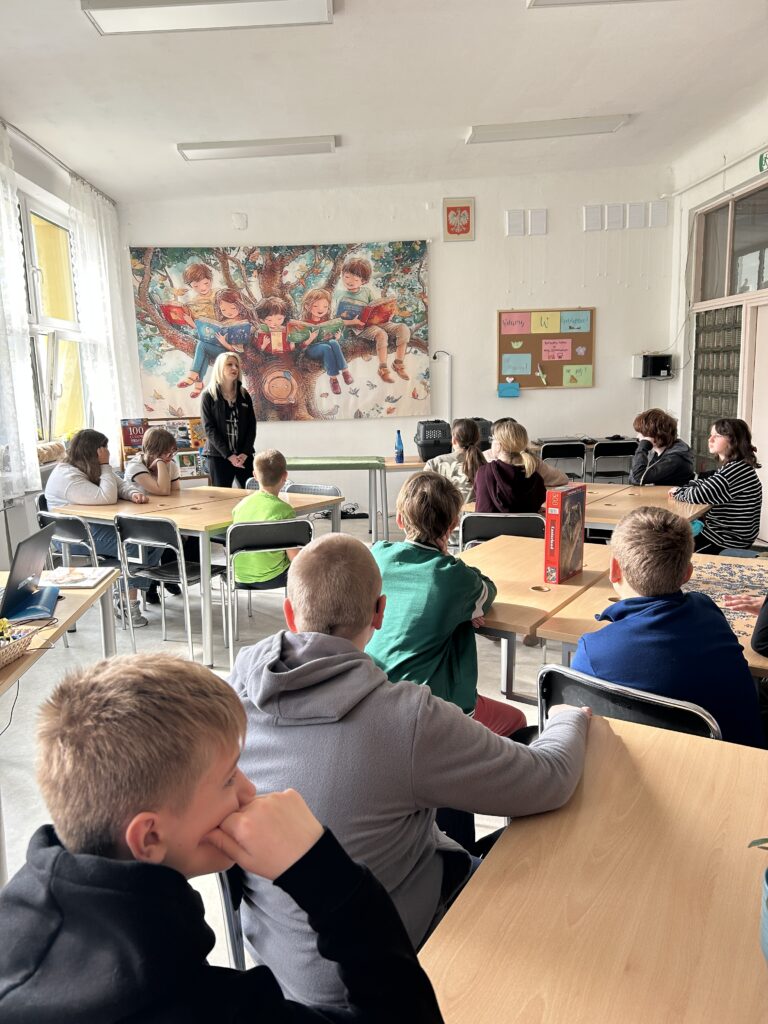w bibliotece na tle baneru z dziećmi siedzącymi na drzewie z książkami stoi pani i opowiada . Przed nią przy stołach siedzą dzieci i słuchają prelekcji o zwierzętach