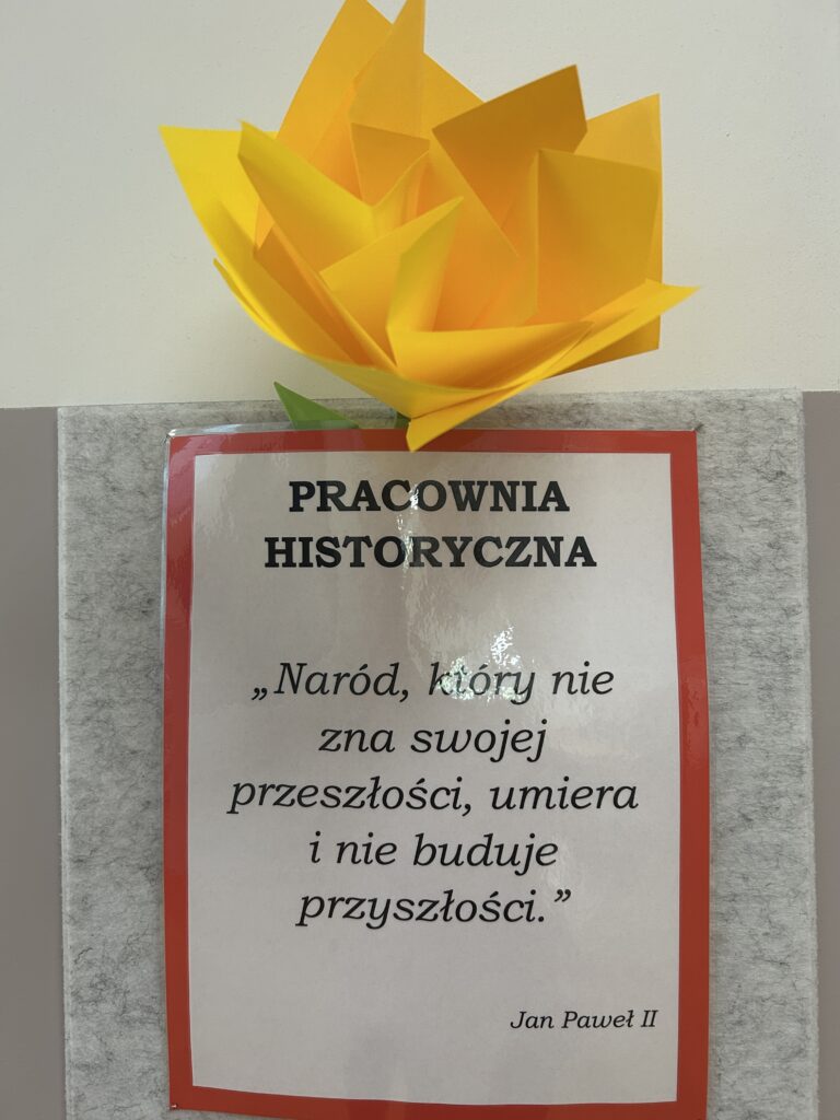 kartka przy pracowni historycznej ze słowami: Pracownia historyczna oraz motto:Naród, który nie zna swojej przeszłości, umiera i nie buduje przyszłości Jan Paweł II. Za tabliczkę wsunięty żóły kwiat zpapieru