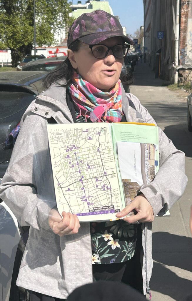 nauczycielka stoi przodem do zdjęcia, w ręku trzyma książkę, która pokazuje dzieciom. W książce jest plan Łodzi z zaznaczonymi muralami oraz zdjęcia murali