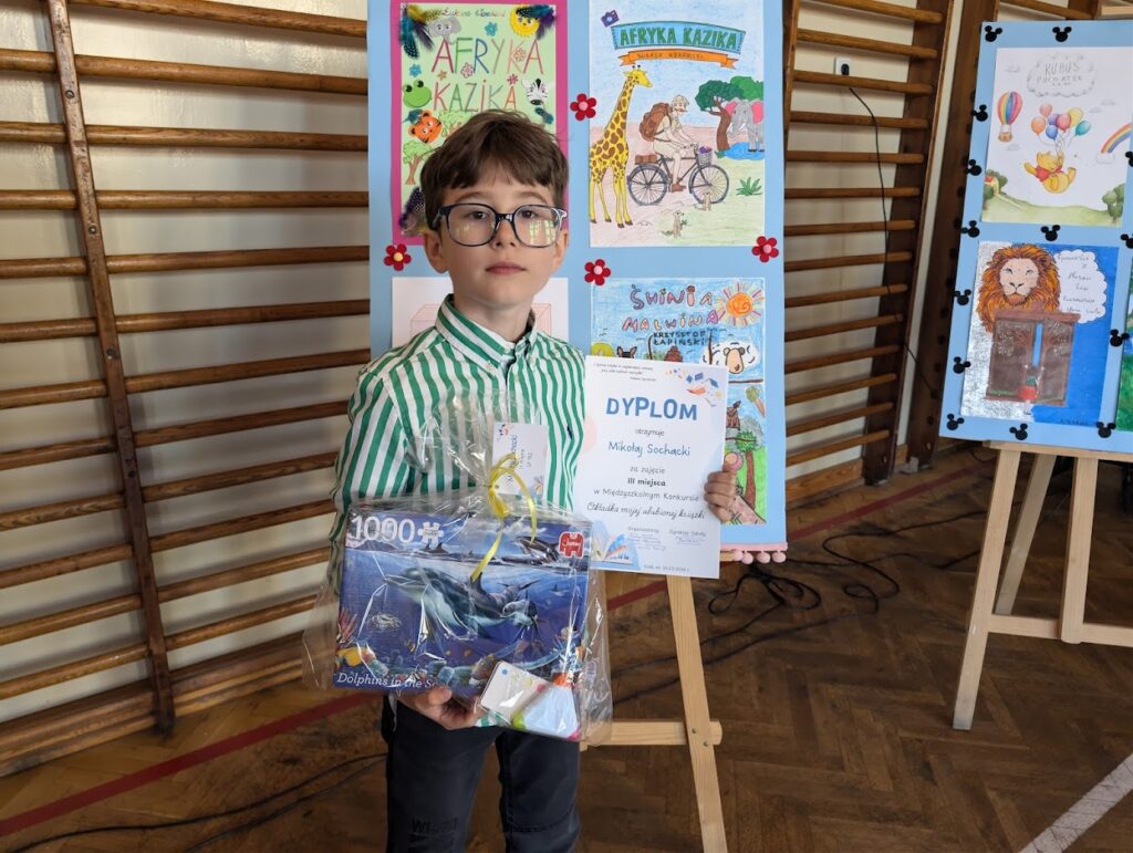 uczeń trzyma dyplom  i nagrodę, stoi przy swojej pracy 