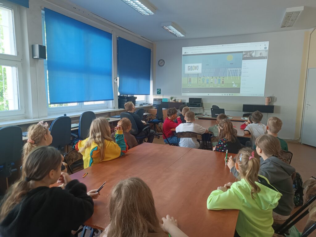 dzieci siedzą w ławkach i patrzą na ścianę z wyświetlonym ekranem