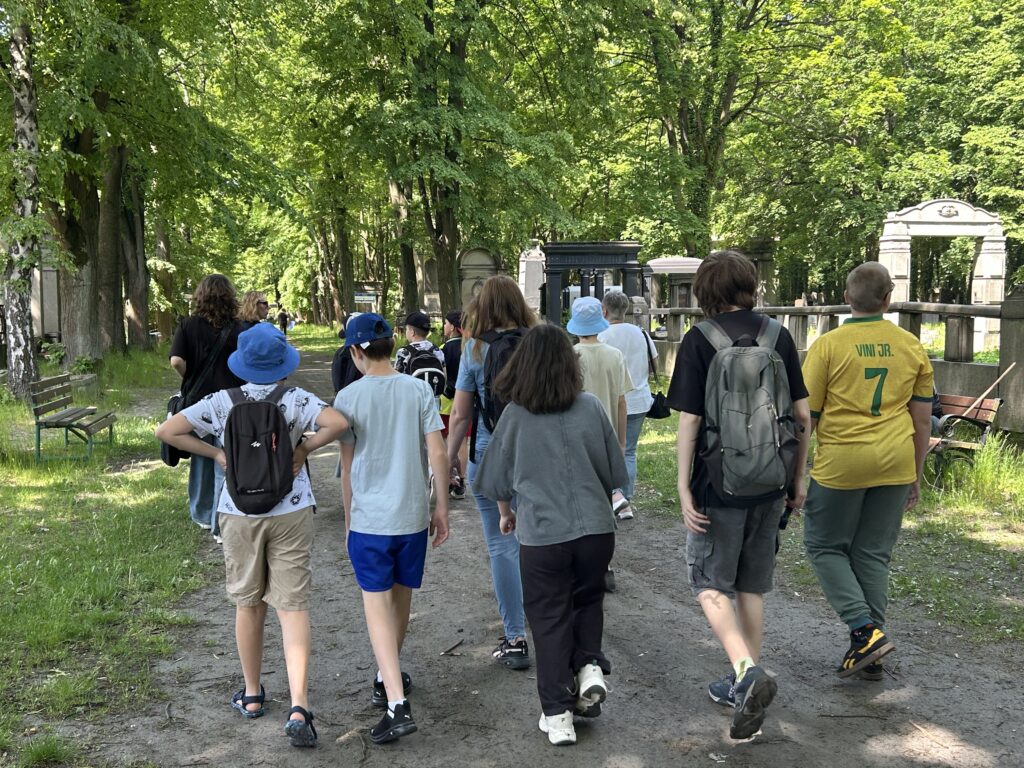 dzieci idą za panią przewodnik główną aleją cmentarną i oglądają nagrobki, które znajdują się po lewej i prawej stronie