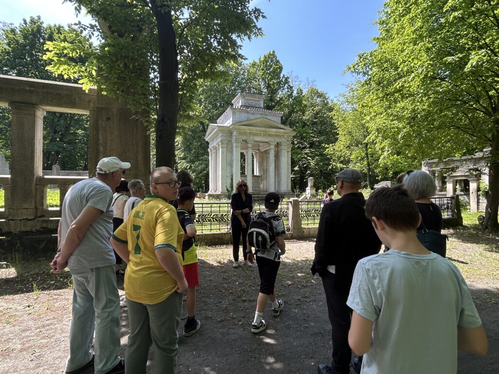 młodzież i dorośli stoją przodem do pani przewodnik, tyłem do aparatu. Przed zebranym grobowiec rodziny Silbersteinów