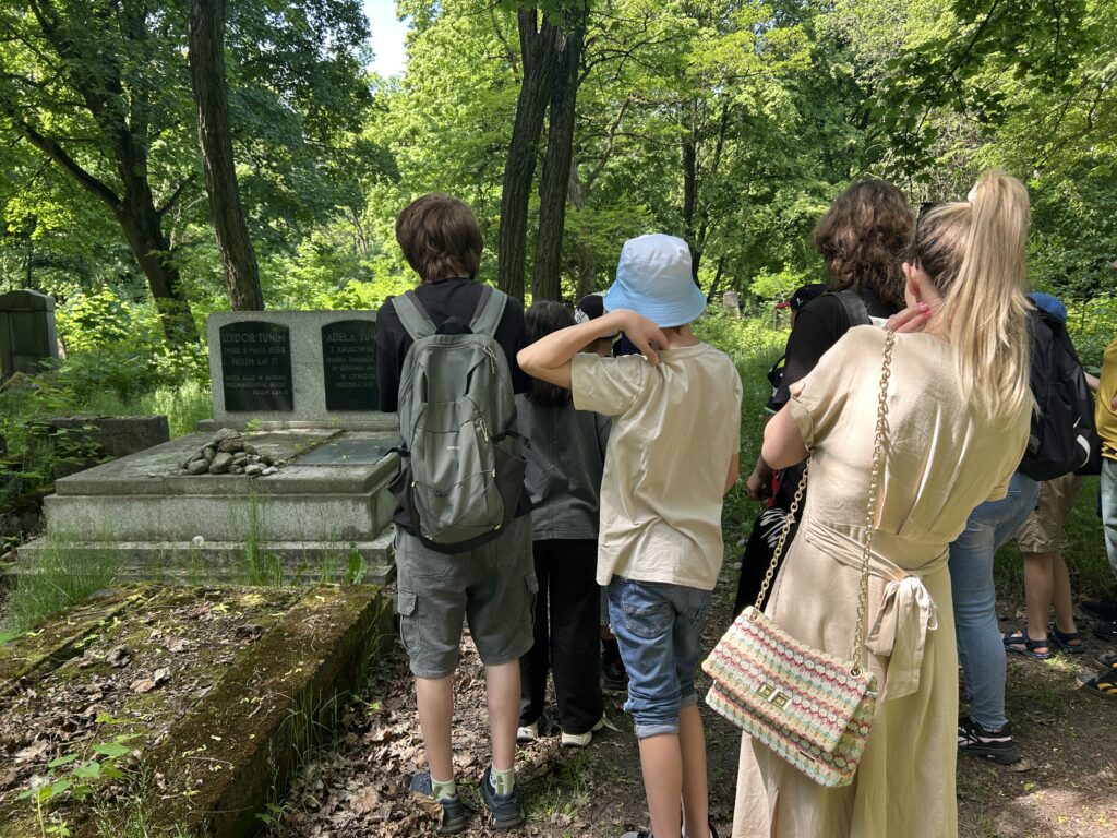pani przewodnik opowiada o rodzicach Juliana Tuwima. Dzieci i dorośli oglądają grób. Na ostatnim zdjęciu sam pomnik rodziców Tuwima z napisami: Izydor Tuwim, Adela Tuwim i tabliczka: Tu spoczywają BP Rodzice Juliana Tuwima