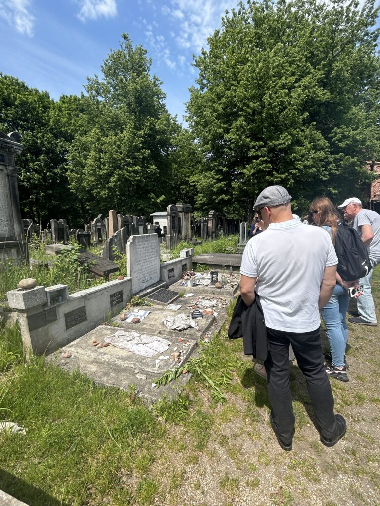 dzieci i dorośli stoją przed grobem na którym leżą koszule oraz kamienie