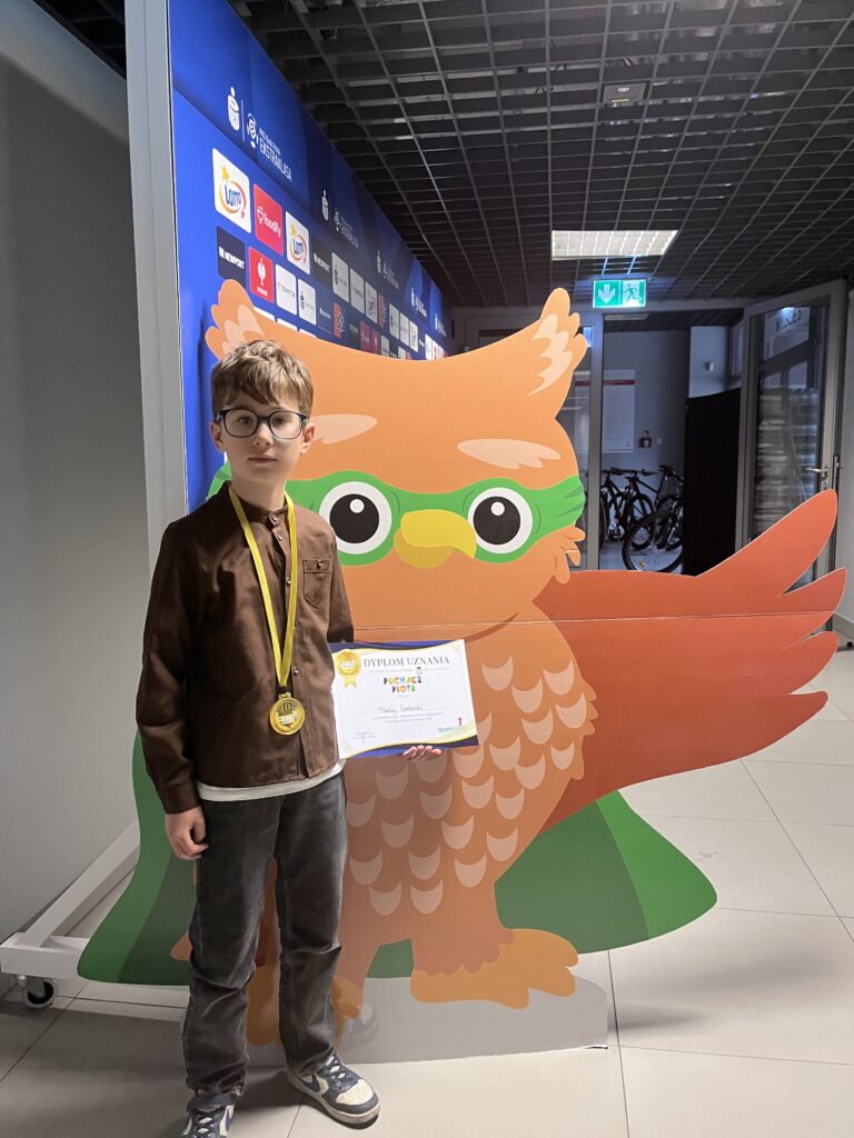 uczeń trzyma dyplom i medal, stoi obok dużego rysunku sowy