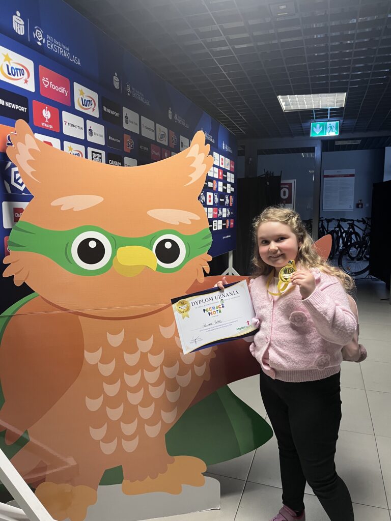 uczennica trzyma dyplom i medal, stoi obok dużego rysunku sowy