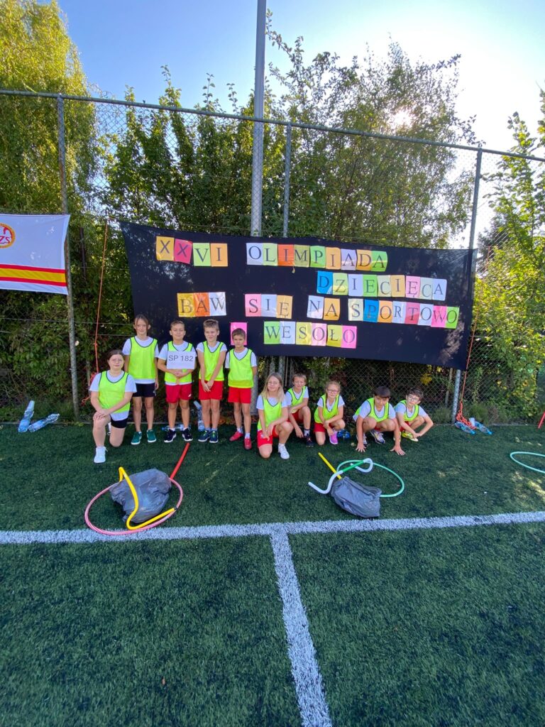 zdjęcie grupowe dzieci przy wielkim plakacie z kolorowym napisem że to 26 olimpiada