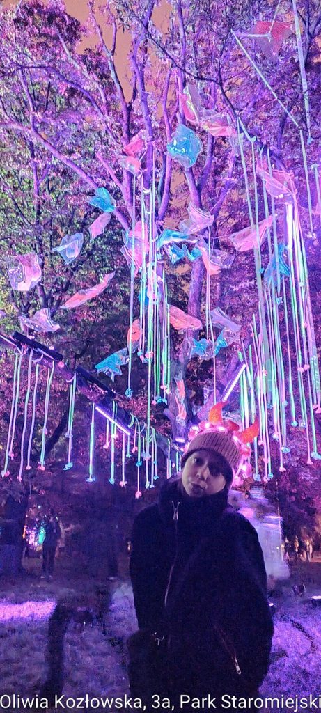 Oliwia Kozłowska - Park Staromiejski - dziewczynka stoi w czapce a na niej świecące rogi, z tyłu instalacja na drzewach wiszące kolorowe sznurki
