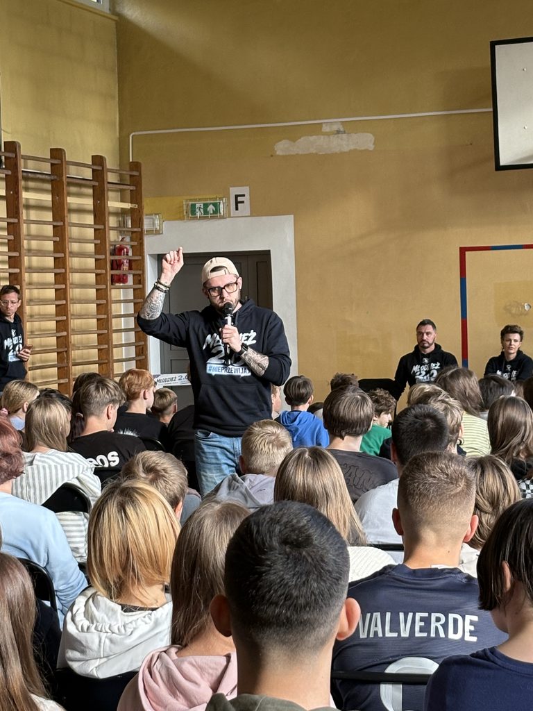 pan Rafał przemawia do młodzieży siedzącej w sali gimnastycznej